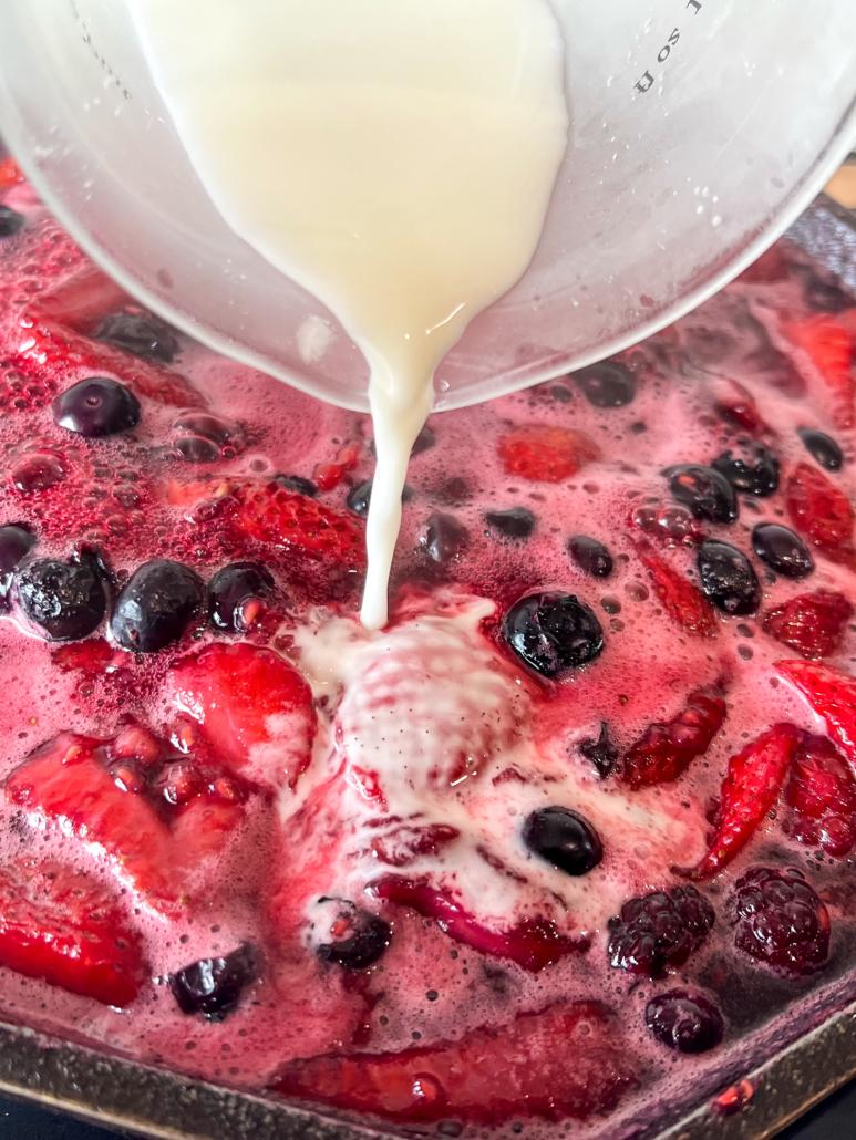 A cornstarch slurry is being poured into the skillet of simmering mixed fresh berries.