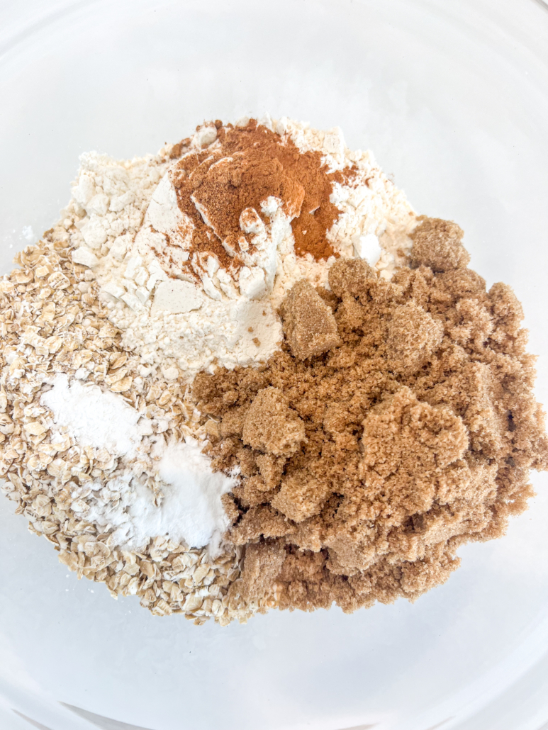 Oats, flour, brown sugar, cinnamon, nutmeg, baking soda, and baking powder in a glass bowl.