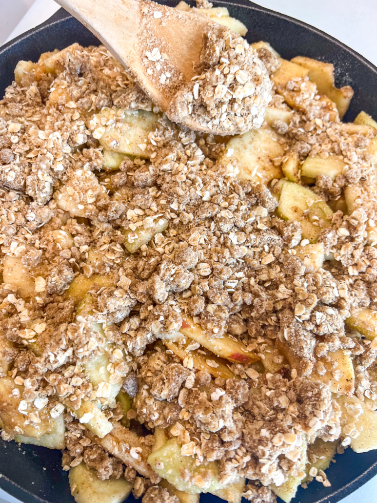 Oat crisp topping being spooned over the seasoned apple pieces in the cast iron skillet.