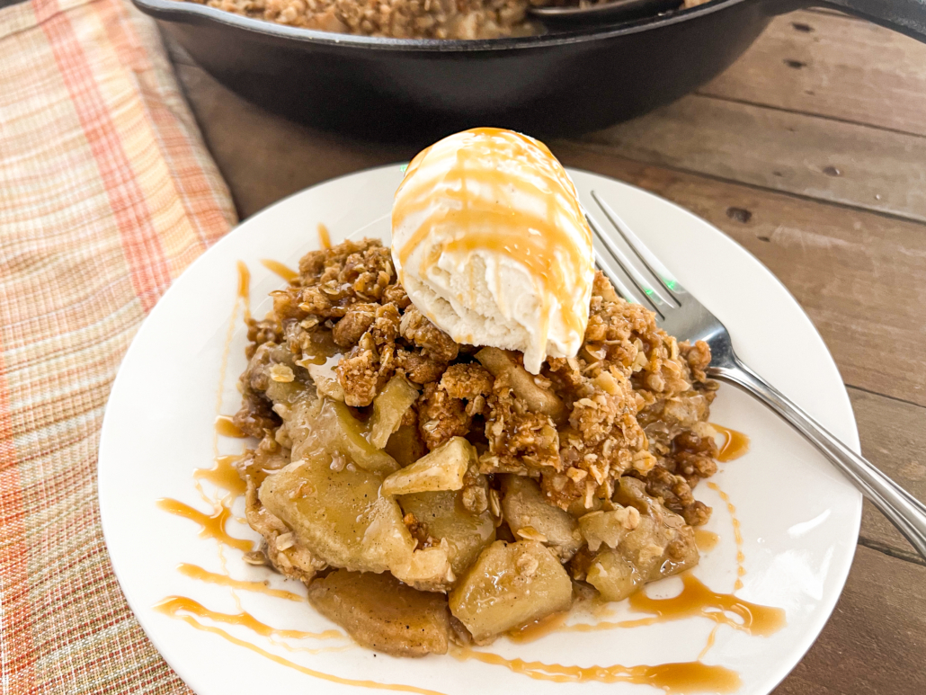 A serving of apple crisp with the skillet in the background topped with vanilla ice cream and caramel drizzle.