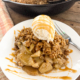 A serving of apple crisp with the skillet in the background topped with vanilla ice cream and caramel drizzle.