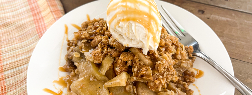 A serving of apple crisp with the skillet in the background topped with vanilla ice cream and caramel drizzle.