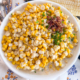Creamy corn in a white bowl garnished with bacon crumbles and chives with spoons and fresh corn cobs in the background.