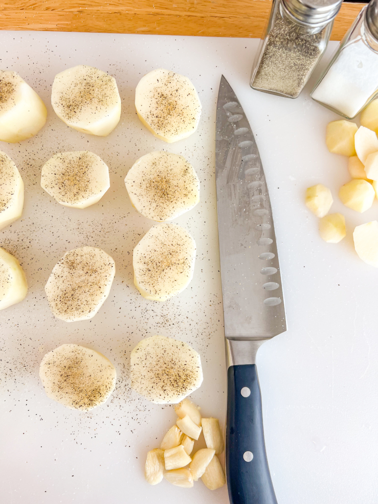 Potatoes have been sliced into one-inch discs, flat on both sides, and sprinkled with salt and pepper, plus roughly chopped garlic on the cutting board with a knife.