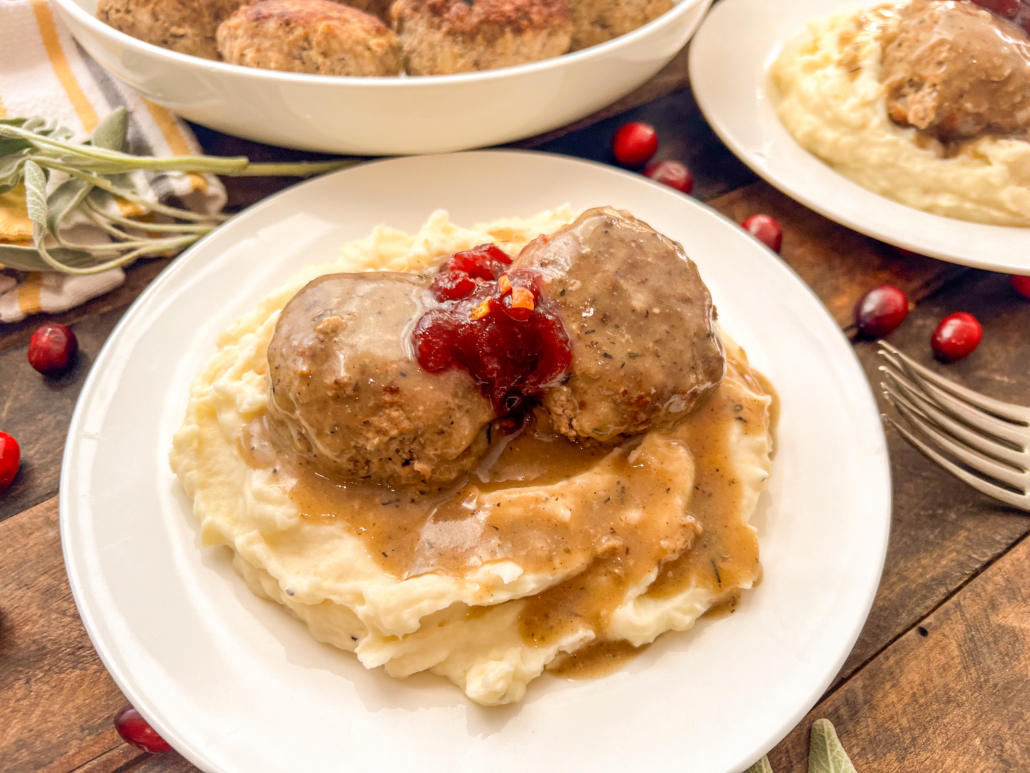 Two turkey and stuffing meatballs served over mashed potatoes with gravy and a dollop of cranberry sauce on top.