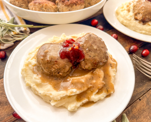 Two turkey and stuffing meatballs served over mashed potatoes with gravy and a dollop of cranberry sauce on top.