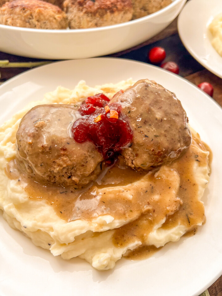 Two turkey and stuffing meatballs served over mashed potatoes with gravy and a dollop of cranberry sauce on top.