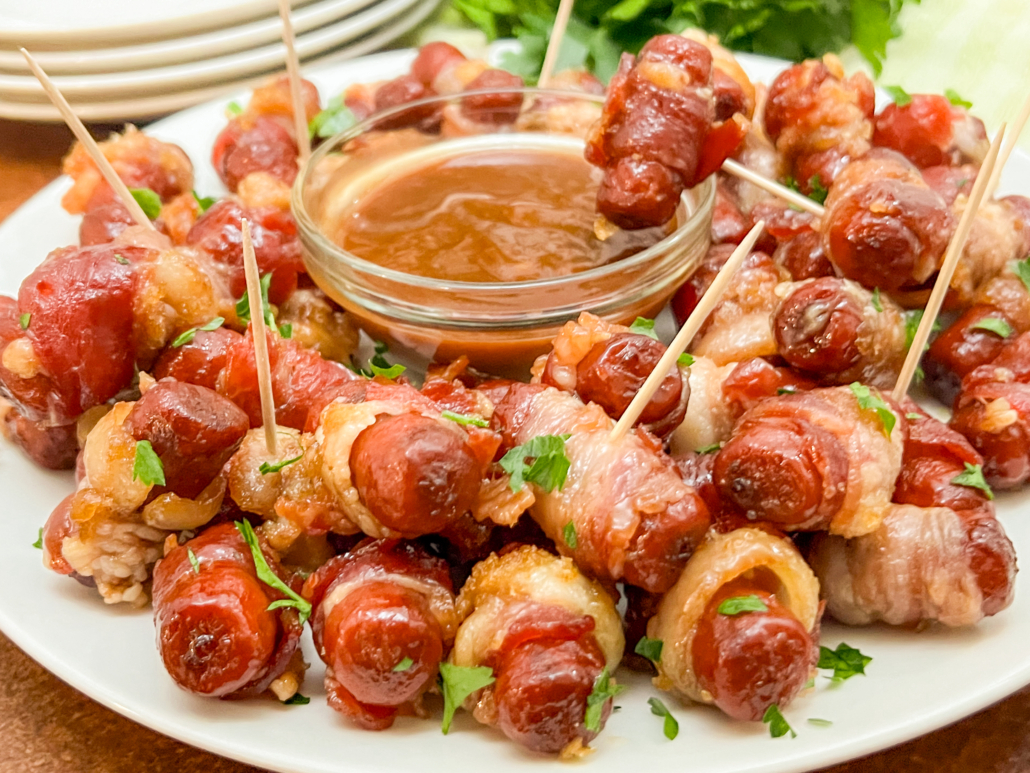 A serving plate of brown sugar coated, bacon wrapped mini sausages and a bowl of barbecue sauce for dipping.