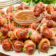 A serving plate of brown sugar coated, bacon wrapped mini sausages and a bowl of barbecue sauce for dipping.