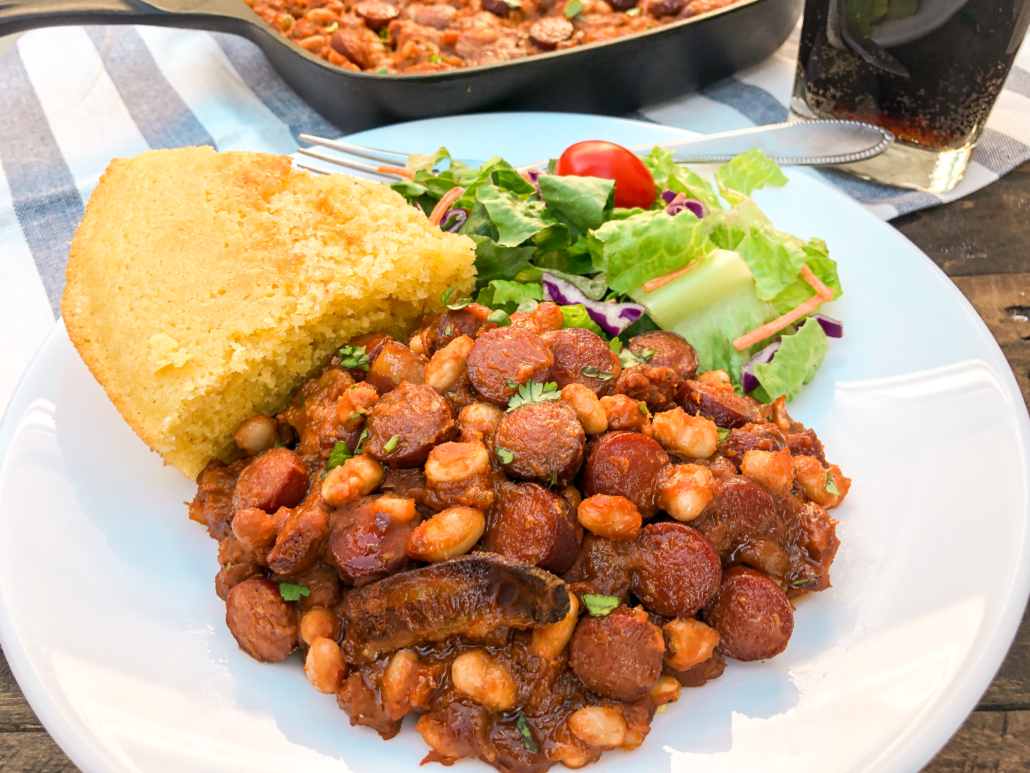 A serving of beans and franks with a piece of cornbread and a green salad on a white plate.