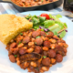 A serving of beans and franks with a piece of cornbread and a green salad on a white plate.