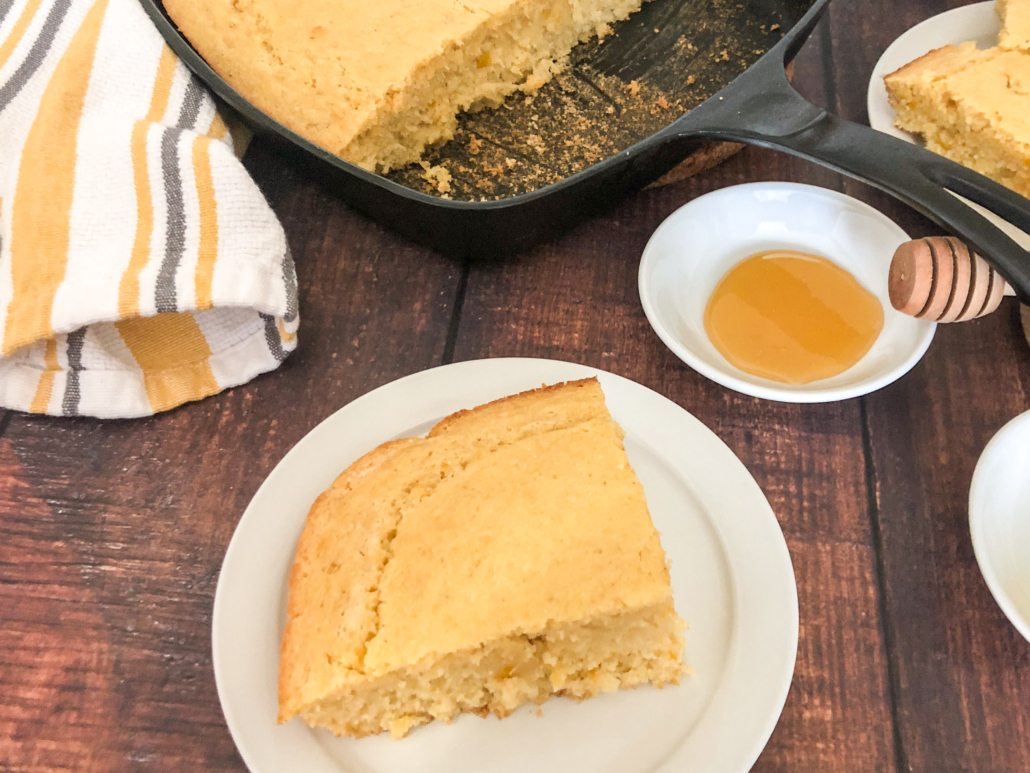 A piece of cornbread on a white plate with the remaining left in the skillet.