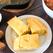 3 pieces of cornbread on a white plate with a bowl of chili and the skillet in the background.