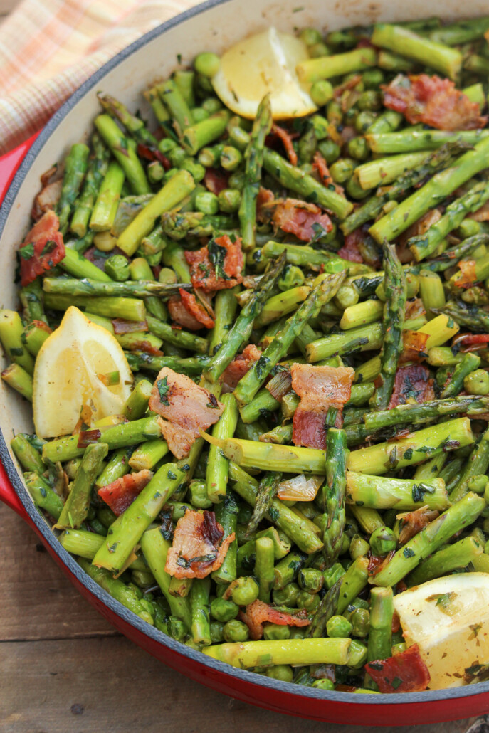 Close up shot of asparagus and peas tossed with bacon and herbs with half-cut lemon wedges as garnish in a red enameled cast iron pan.