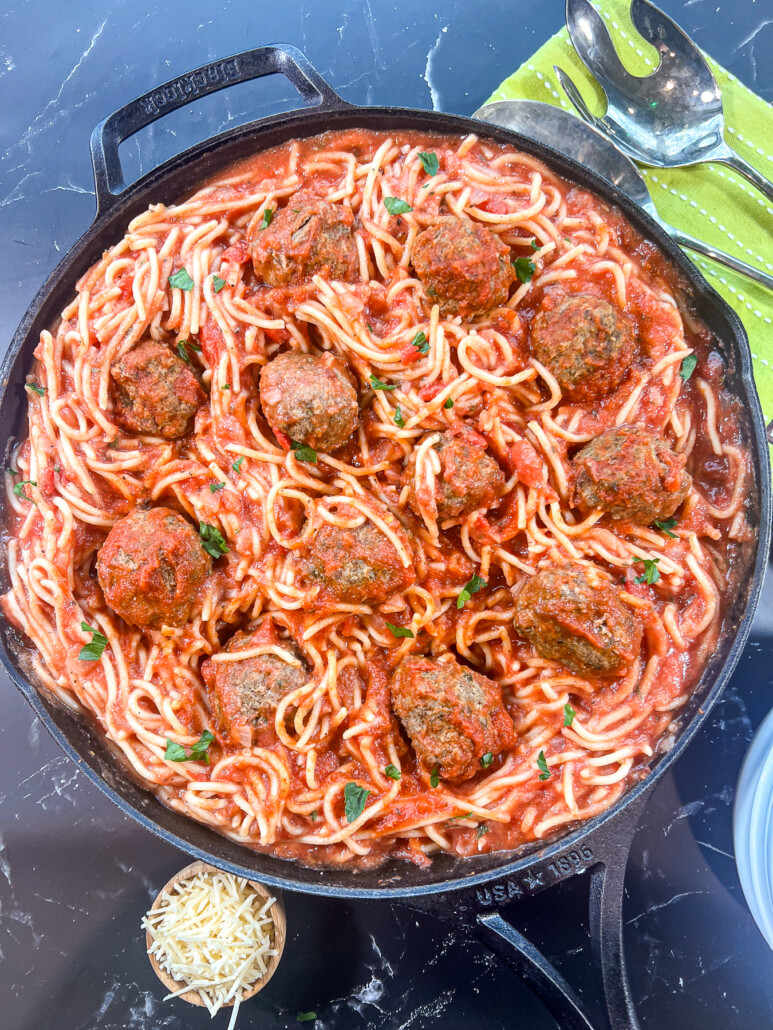 Spaghetti and Meatballs in a cast iron skillet garnished with fresh parsley and a small bowl of parmesan cheese for garnishing.