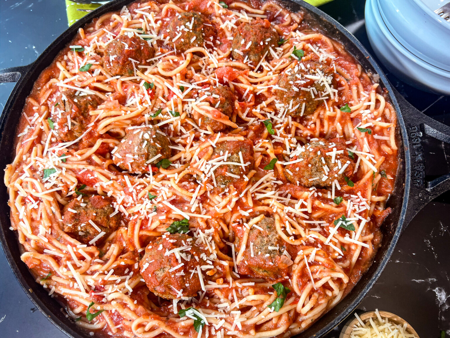 A cast iron skillet filled with spaghetti and meatballs topped with shredded cheese and fresh herbs in a rich tomato sauce.