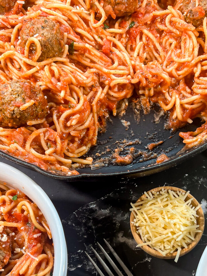 Close up image of the skillet with spaghetti and meatballs, two portions missing and plated in white bowls.