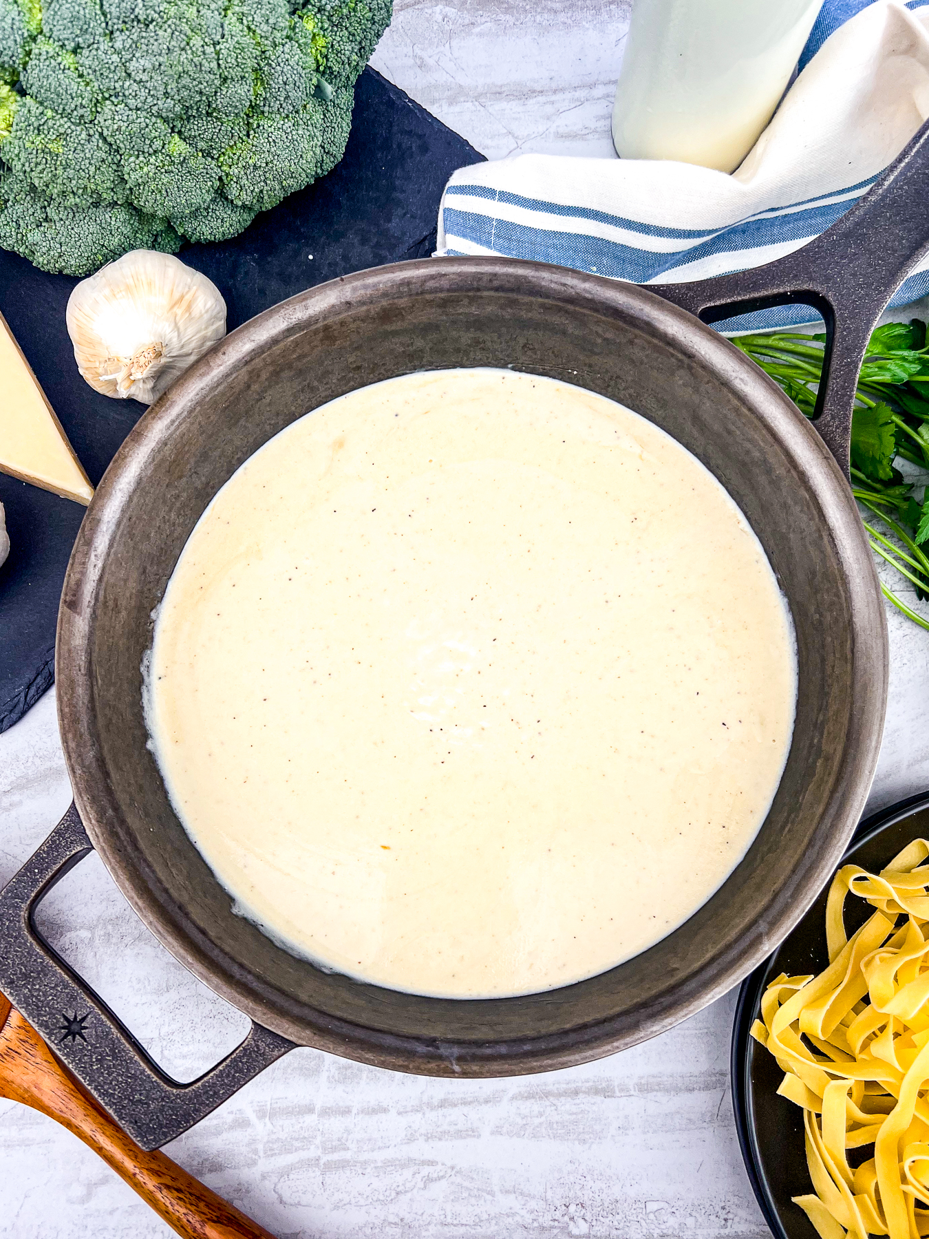 Easy creamy homemade alfredo sauce in a cast iron skillet with various ingredients to make pasta around the skillet of sauce.