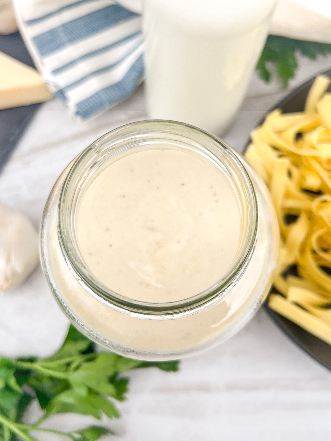Homemade Alfredo sauce in a glass jar with out of focus pasta cream, garlic, Parmesan cheese, and parsley encircling it.