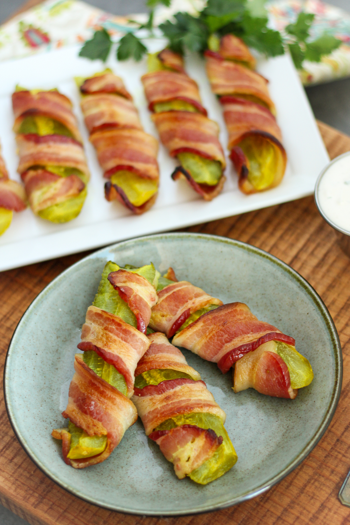A white platter of pickles wrapped in bacon and some on a blue-stoned hued plate.