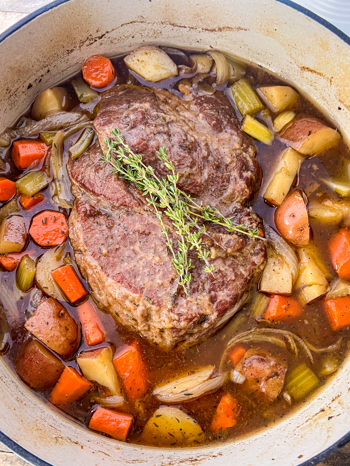 Dutch oven beef roast with carrots, onions, potatoes, and celery sitting in broth and garnished with sprigs of fresh thyme.