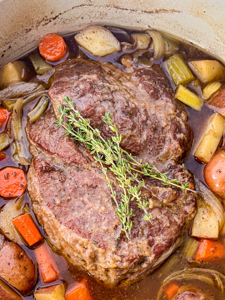Dutch oven beef roast with carrots, onions, potatoes, and onions sitting in broth and garnished with sprigs for fresh thyme.