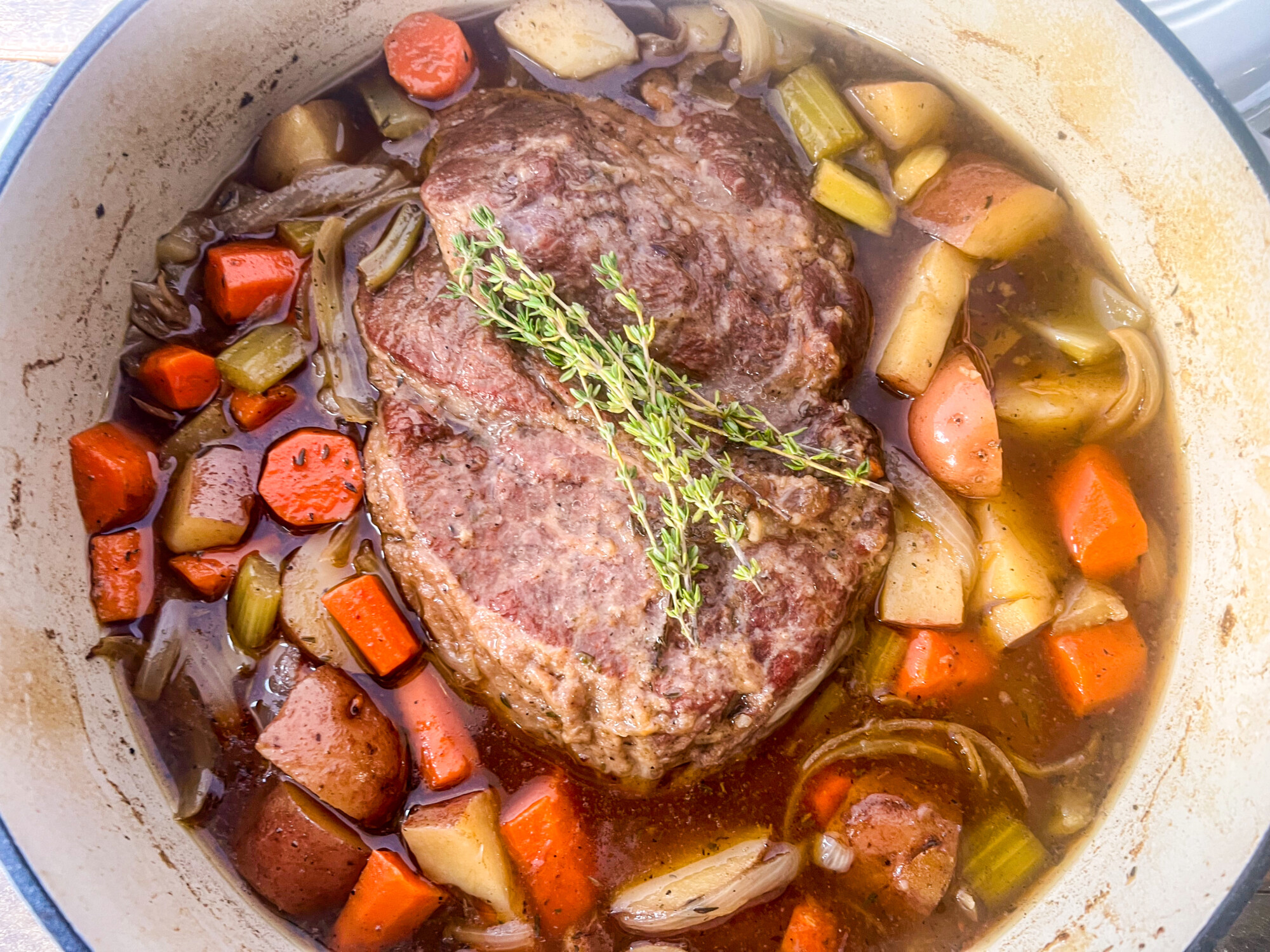 Dutch oven beef roast with carrots, onions, potatoes, and onions sitting in broth and garnished with sprigs for fresh thyme.