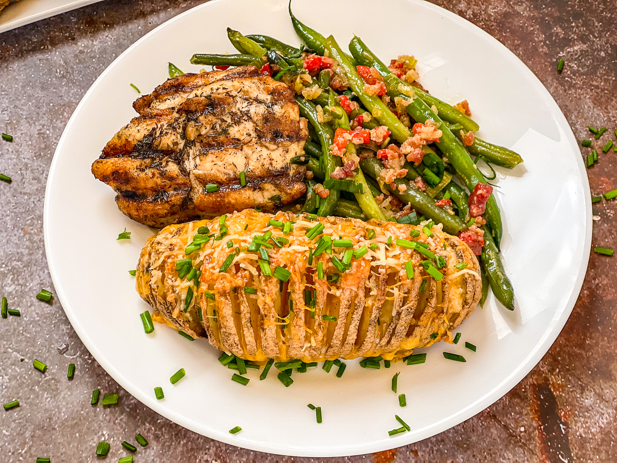 A cheesy hasselback potato garnished with fresh chives on a white plate with grilled chicken and fresh green beans.