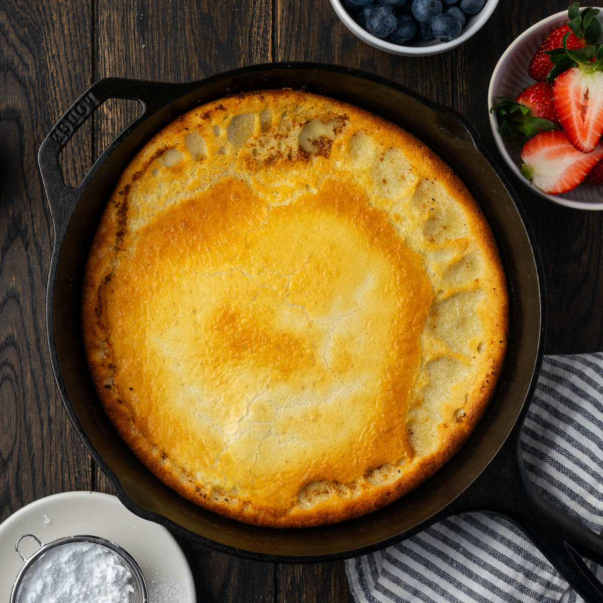 A cooked Dutch baby pancake in a skillet with bowls of fruit and powdered sugar to the side.