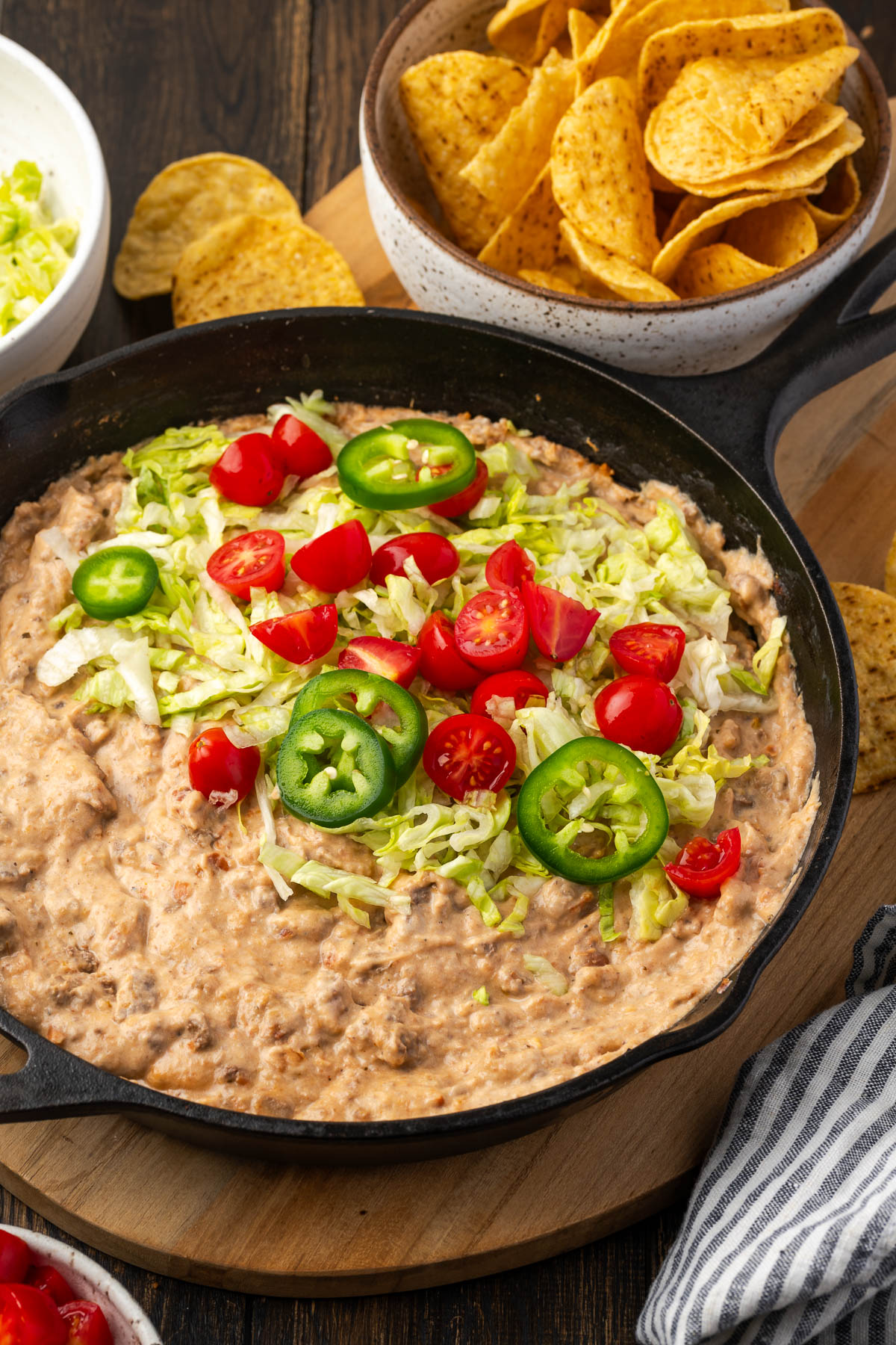 A skillet of nacho bean dip on the table topped with lettuce, jalalpenos, and diced tomato.