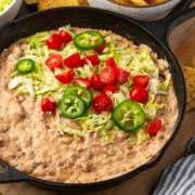 A skillet of nacho dip with lettuce, tomatoes, and jalapenos on top.