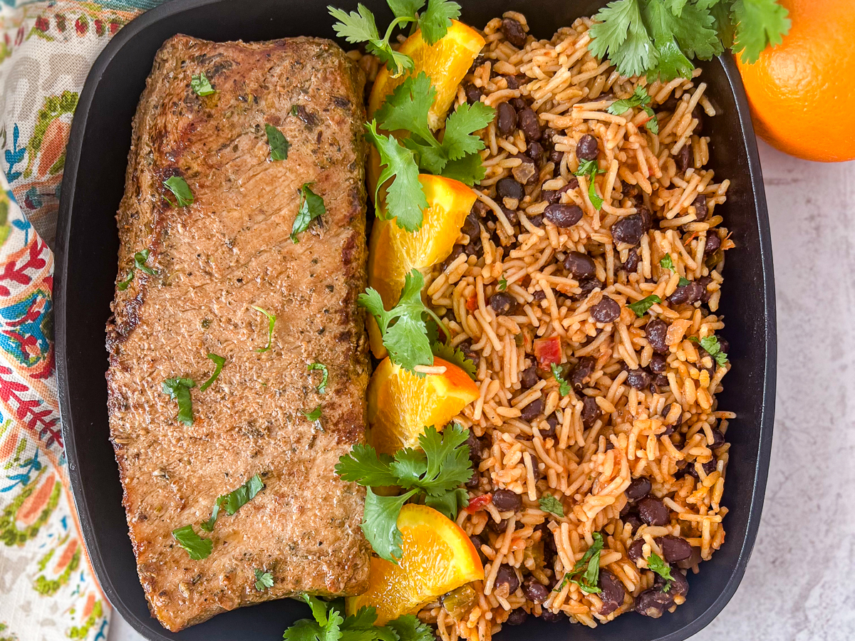Chipotle orange pork tenderloin in a cast iron skillet next to rice and beans with orange and cilantro as garnish.