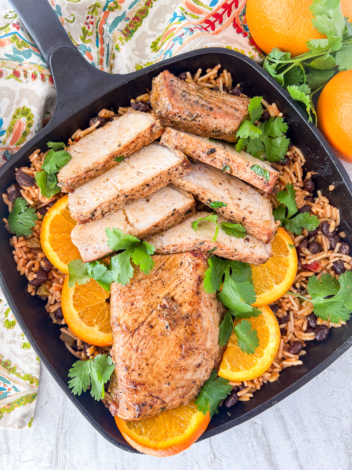 Cast iron skillet with a pork tenderloin half sliced resting on a bed of rice and beans with orange slices and cilantro for garnish.