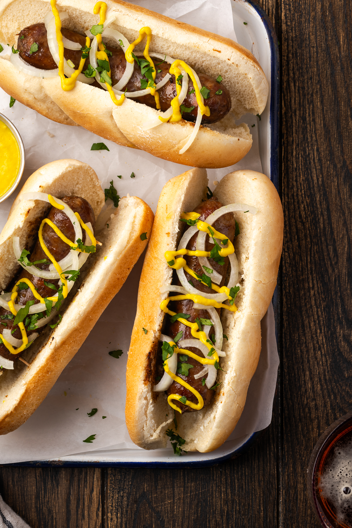 Beer brats on buns with toppings on a baking pan.