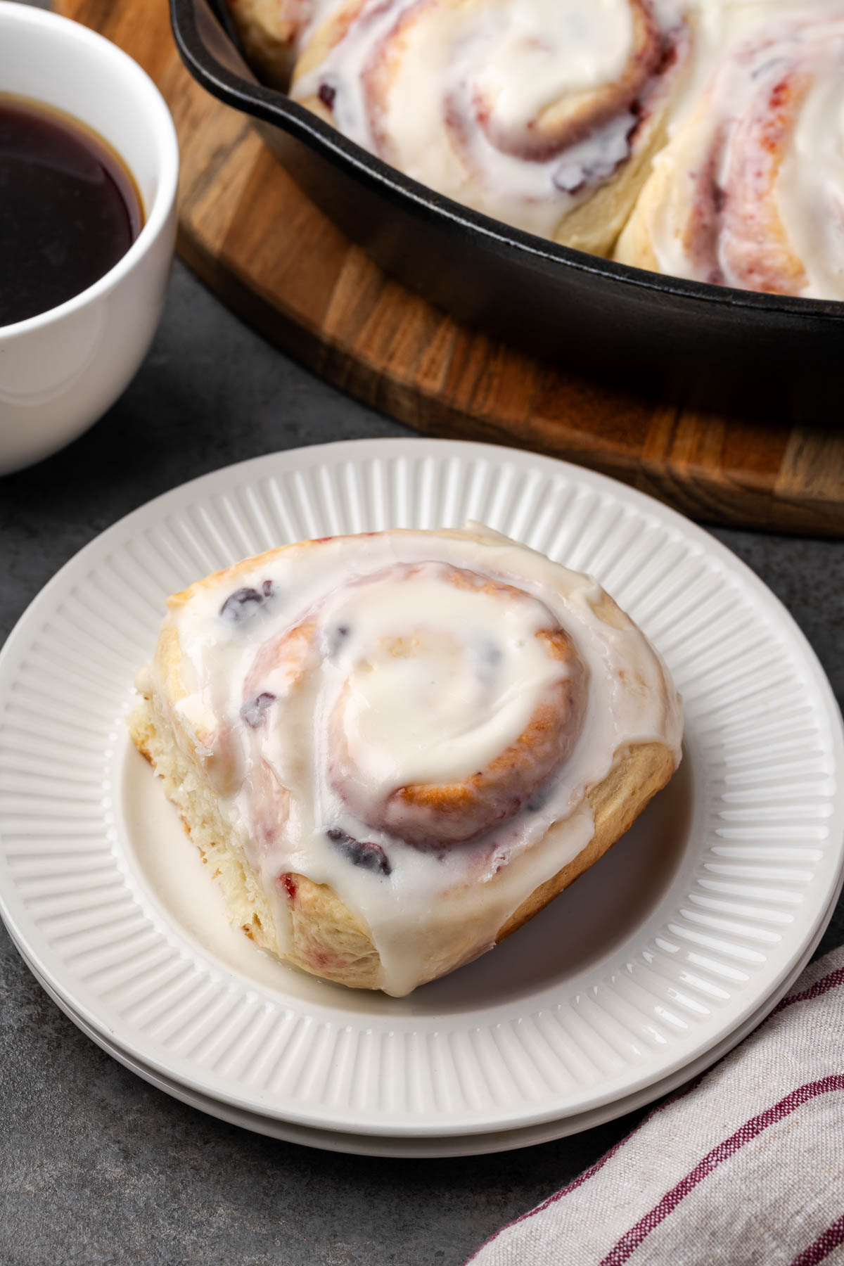 A cherry roll topped with icing on a plate and more in a skillet in the background.