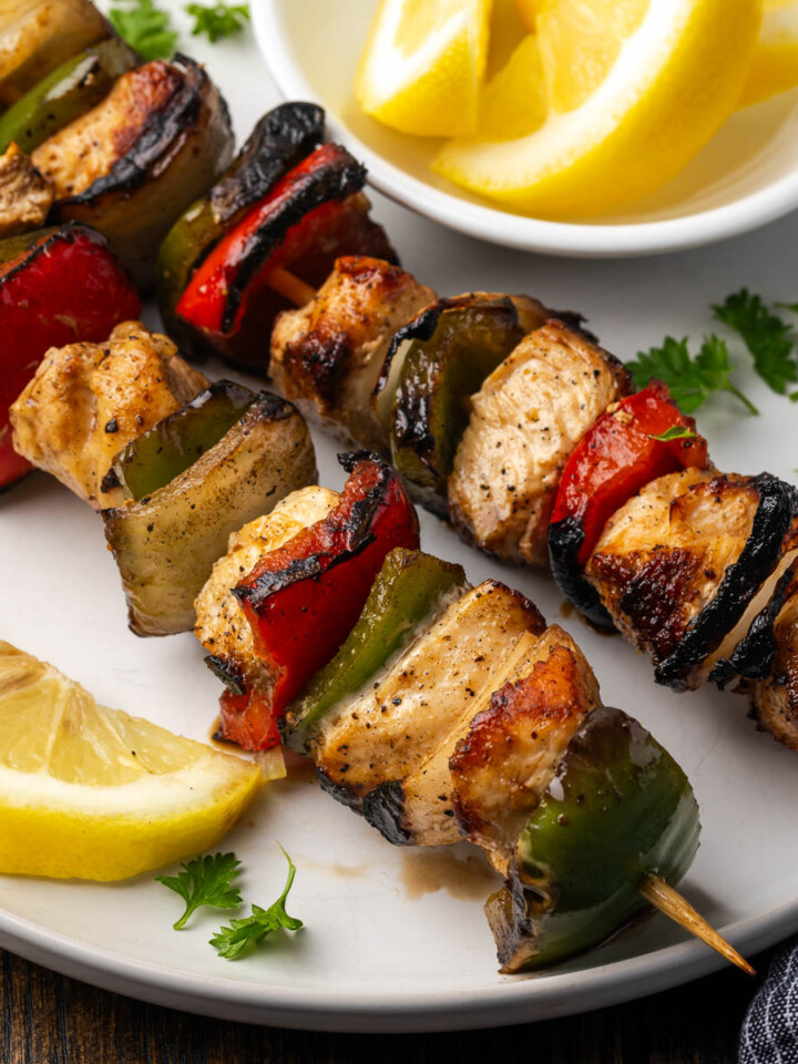 Chicken and veggie kebobs on a plate next to a slice of lemon.