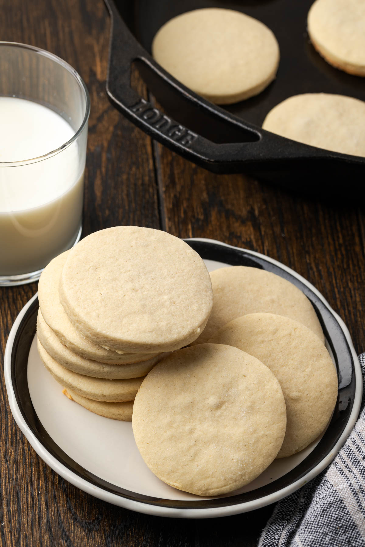 Sugar cookies stacked on a plate next to a glass of milk and more cookies in a cast iron skillet.