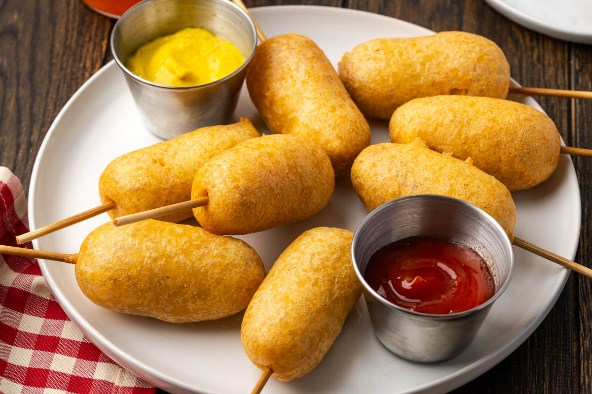 Fried homemade corn dogs on a plate with bowls of ketchup and mustard.