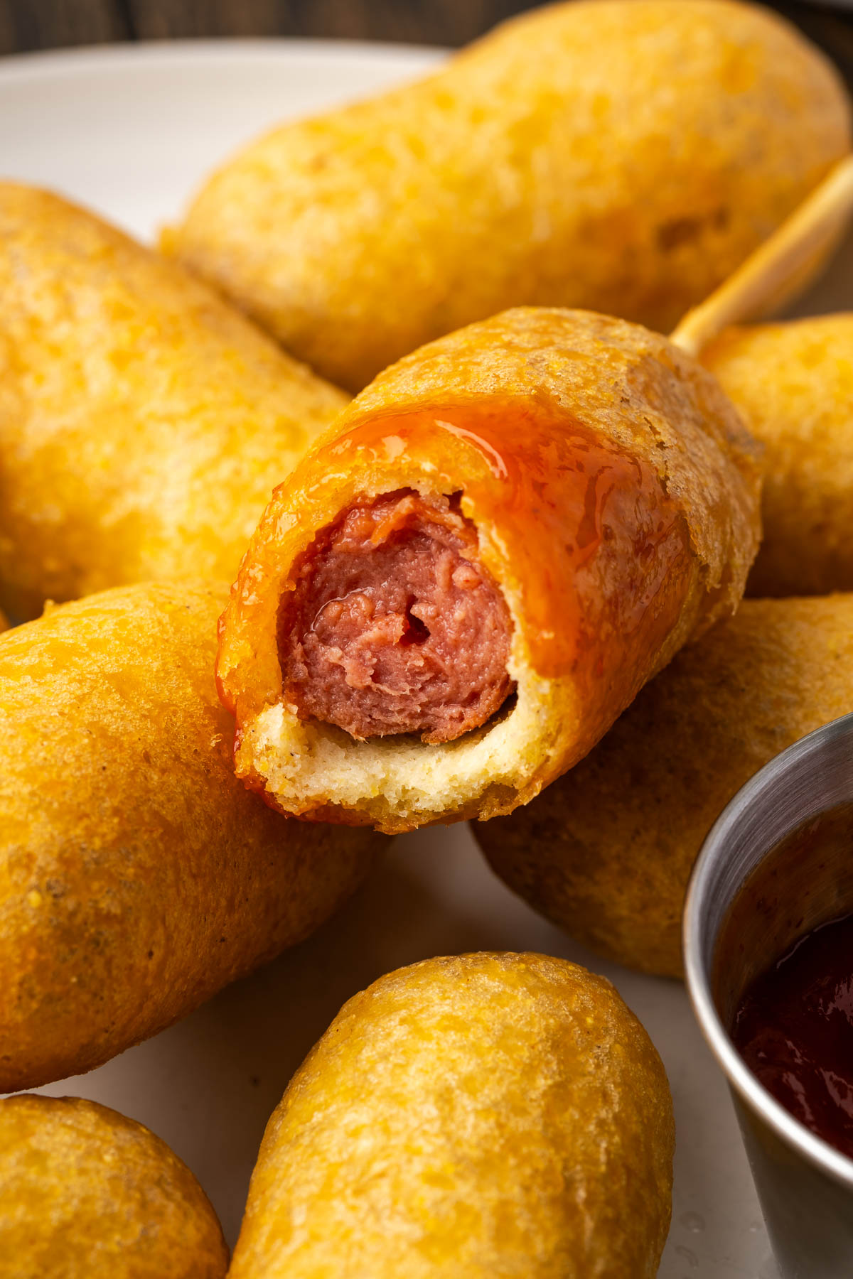 A pile of corn dogs with a bite taken from one with some ketchup on it.