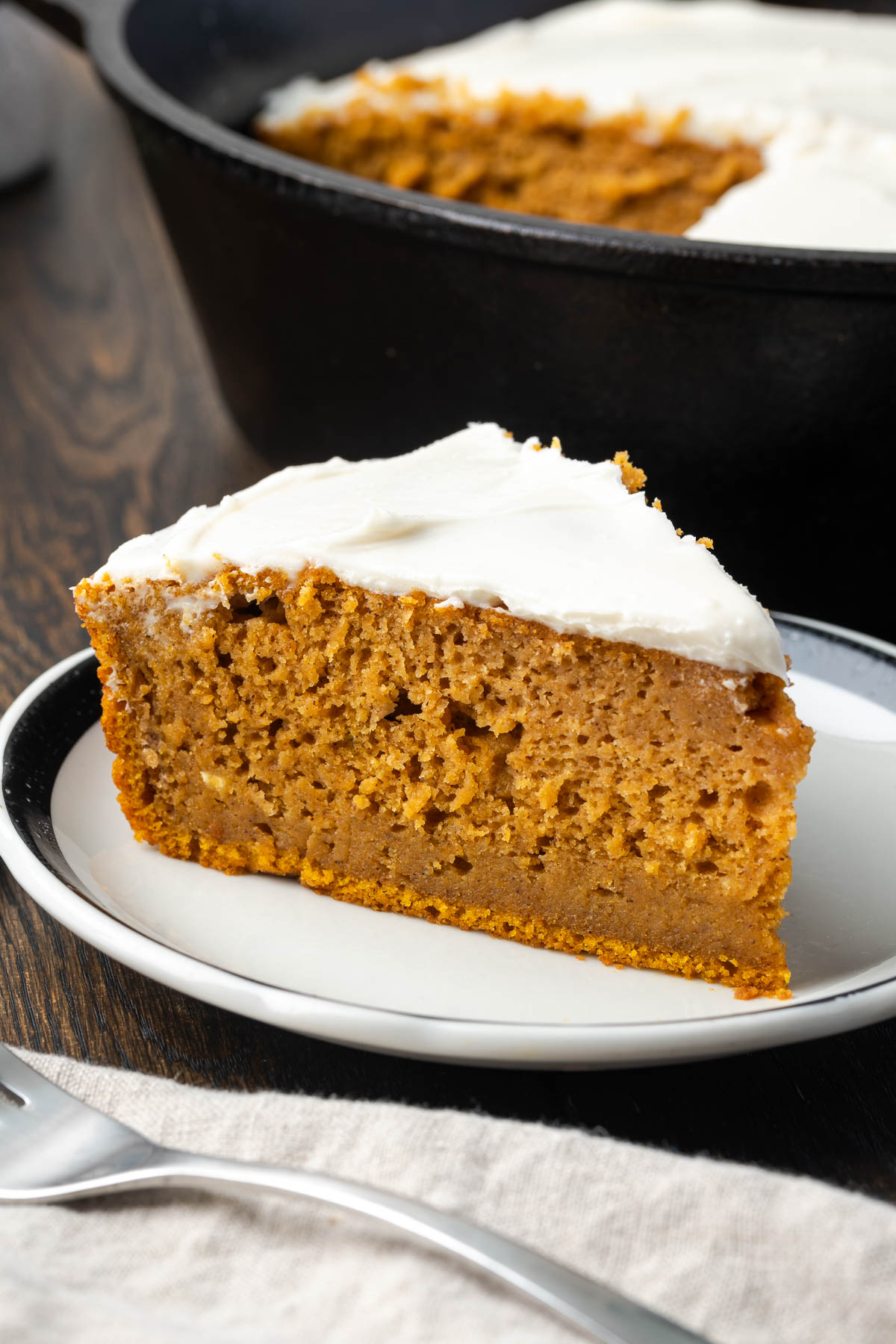 A slice of pumpkin spice cake on a plate with more in a cast iron skillet in the back. 