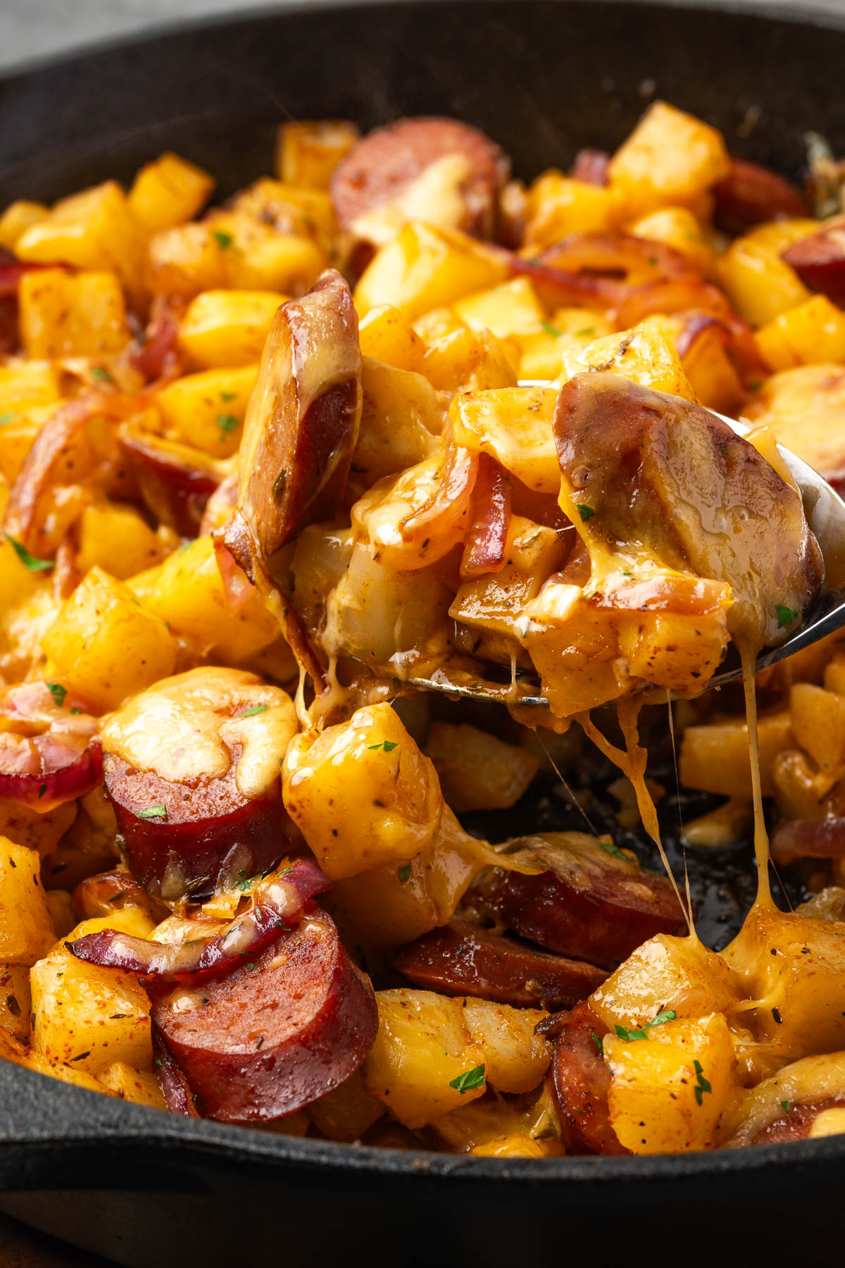 A spoon lifting up a portion of sausage and potatoes from the skillet.
