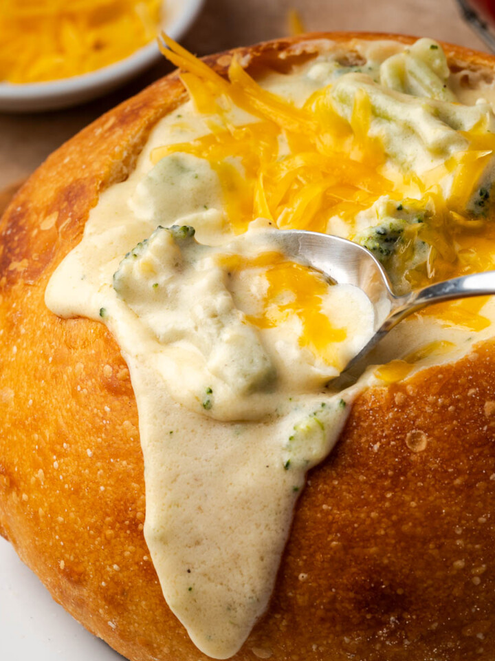 A bowl of broccoli cheese soup in a bread bowl with a spoon dipping some.