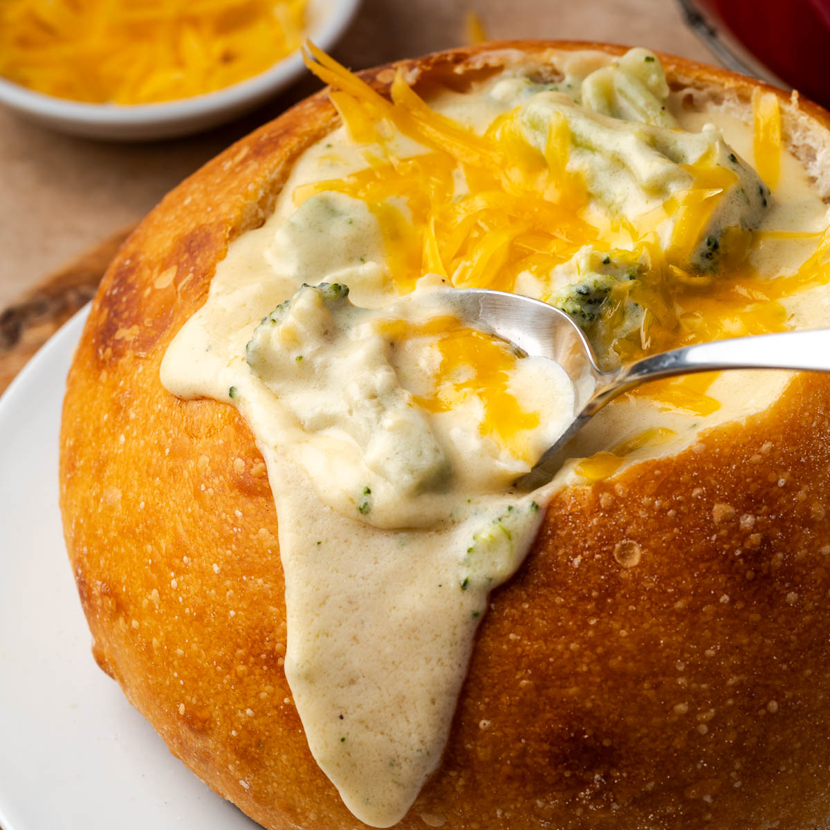 A bowl of broccoli cheese soup in a bread bowl with a spoon dipping some.