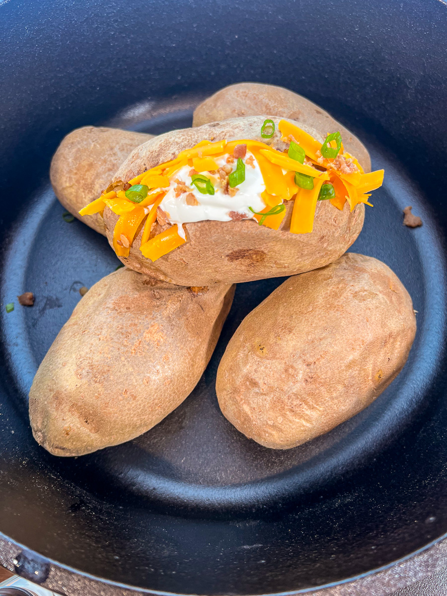 Dutch oven baked potatoes cooked over a campfire in the pot with one on top loaded with toppings.