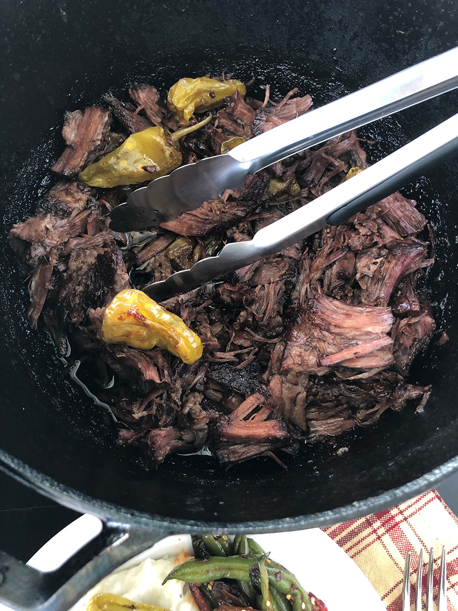 Tongs grabbing shredded Mississippi pot roast from a Dutch oven.