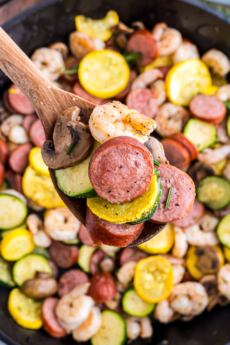 A wooden spoon holding some shrimp, sausage, zucchini, squash up in the air.