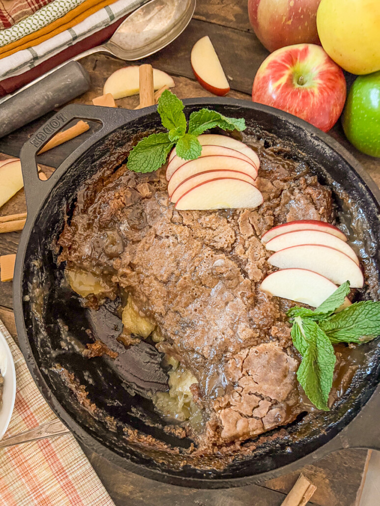 Branded Image of caramel Apple dump cake in a skillet with scoops of ice cream drizzled with caramel sauce, 4 spoons, fresh apple slices, and mint.