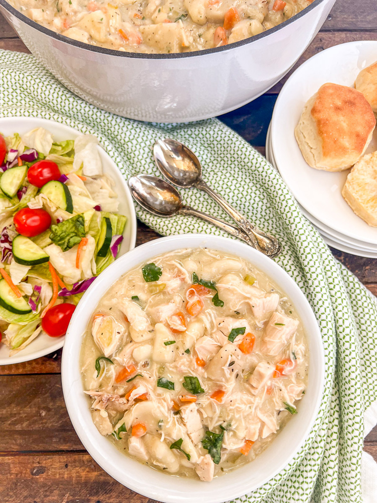 Old fashioned chicken and dumplings in a dutch oven and in a bowl with biscuits and salad as side dishes.