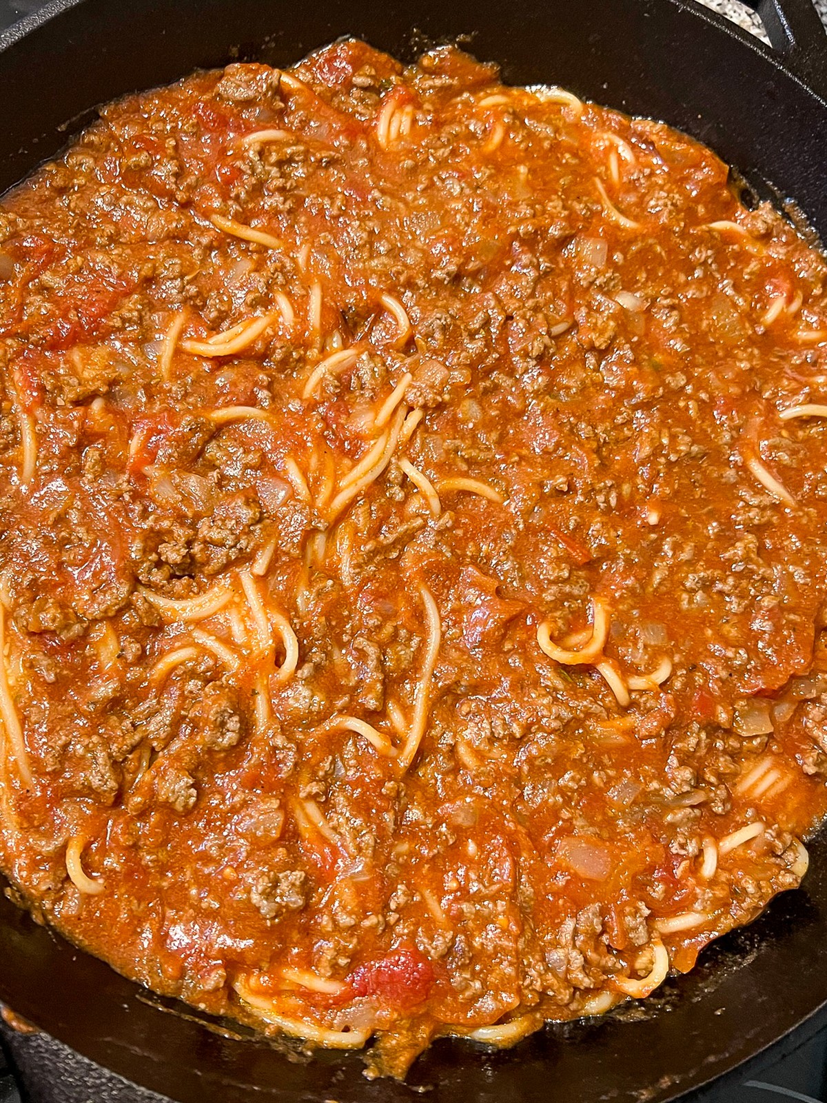 Meat sauce has been evenly spread over the coated spaghetti in the cast iron skillet.
