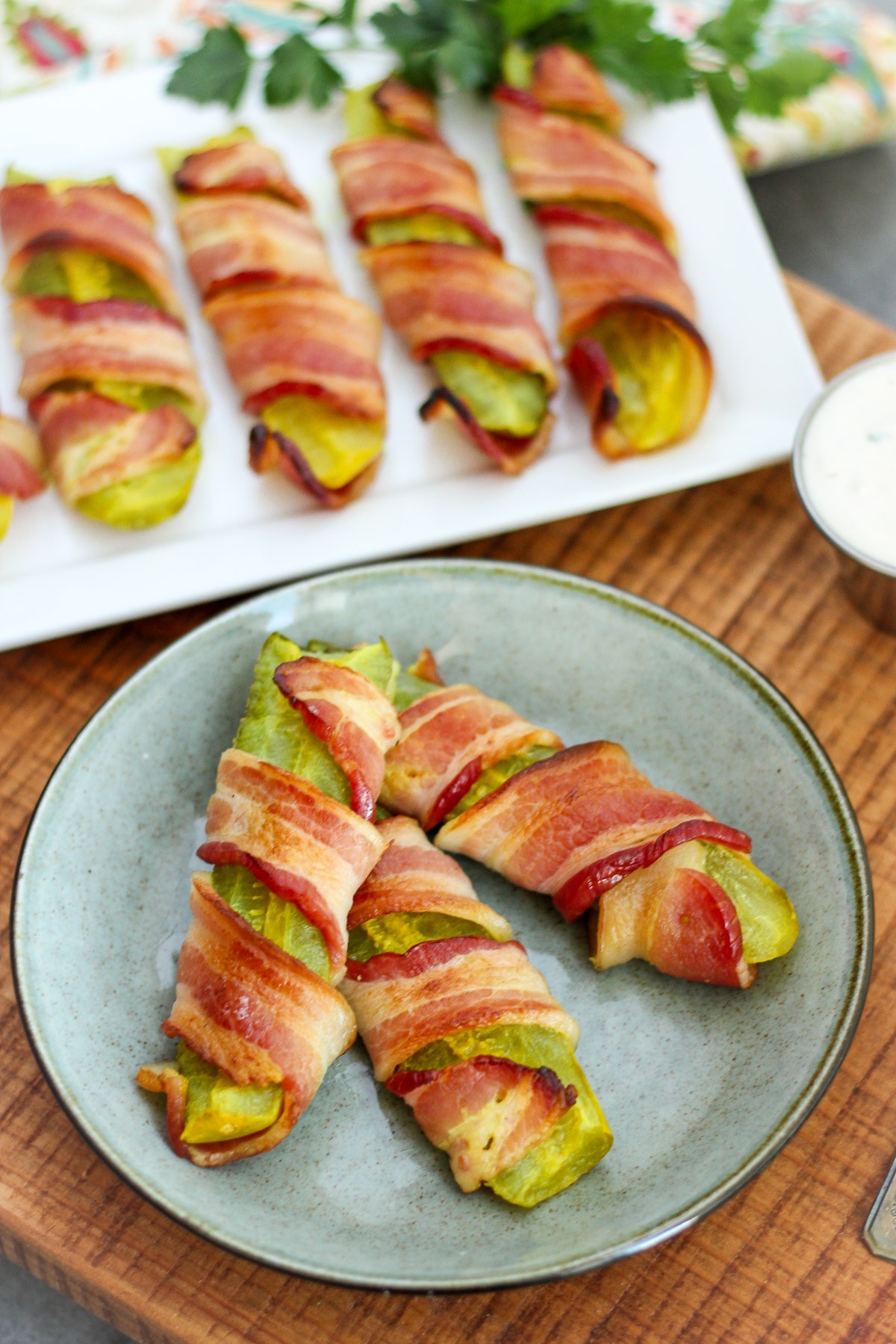 A white platter of pickles wrapped in bacon and some on a blue-stoned hued plate.